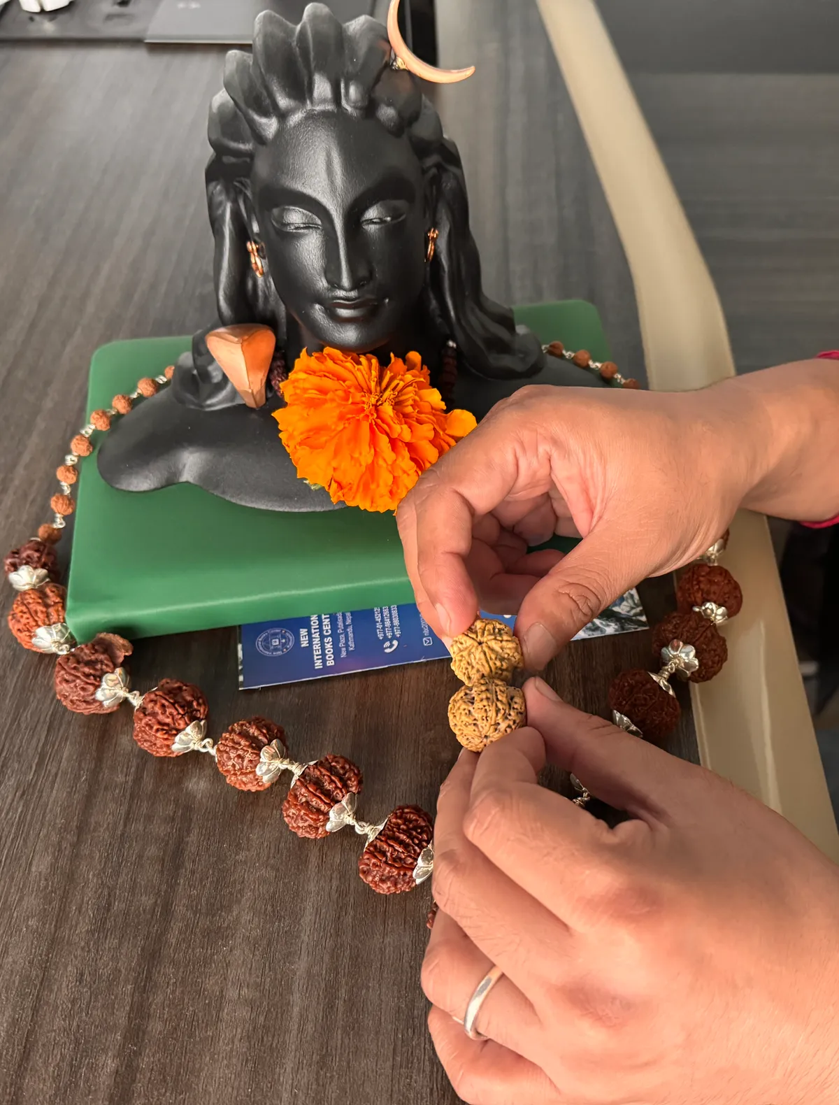 Two pale Rudraksha beads held lightly between fingertips, almost touching, in front of a black Shiva bust and a silver-capped Rudraksha mala