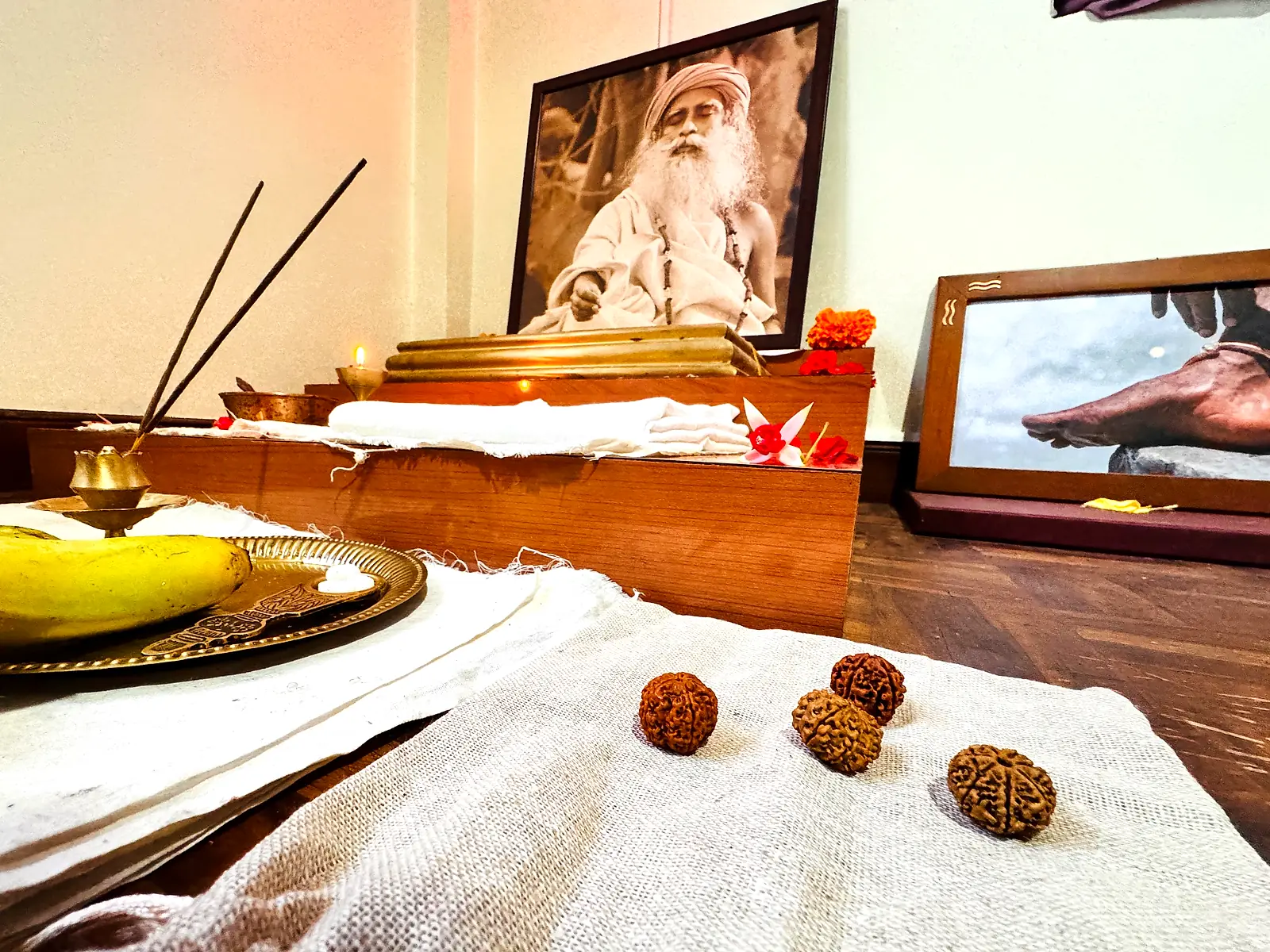 A home altar in Kathmandu at dawn: a lit oil lamp and burning incense before a framed portrait of the guru, marigolds and white cloth on the shrine, and four Nepali rudraksha beads laid out on handwoven cloth in the foreground.